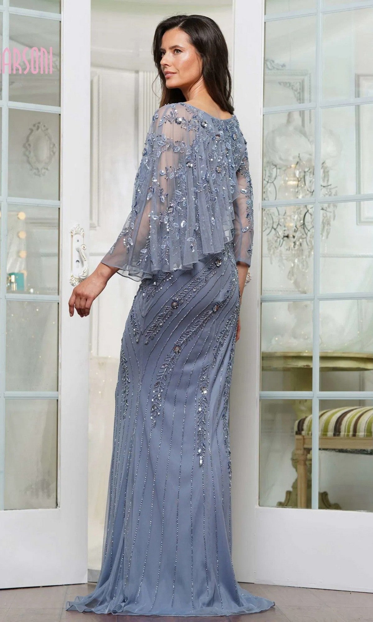 A woman stands indoors wearing the Marsoni MV1343, a floor-length light blue column gown with sequins and a sheer, embellished cape. She faces away, looking to the side.